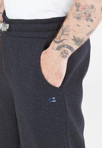Mörkgrå sweatpants i mjukt tyg, med sidofickor och en liten broderad logotyp i blått och vitt nära fållen.