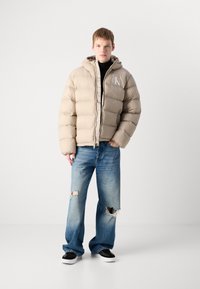 Young man wearing beige puffer jacket with hood, black turtleneck, ripped blue jeans, and black sneakers, standing against a plain white background.