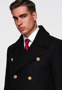 Manteau en laine noire avec un large col, design double boutonnage et boutons dorés. Associé à une chemise blanche et une cravate rouge.