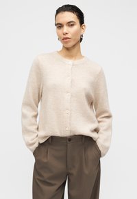 Μπεζ ζακέτα από απαλό πλεκτό ύφασμα, με στρογγυλή λαιμόκοψη και έξι κουμπιά μπροστά. Συνδυάζεται με καφέ παντελόνι ραμμένο στα μέτρα.