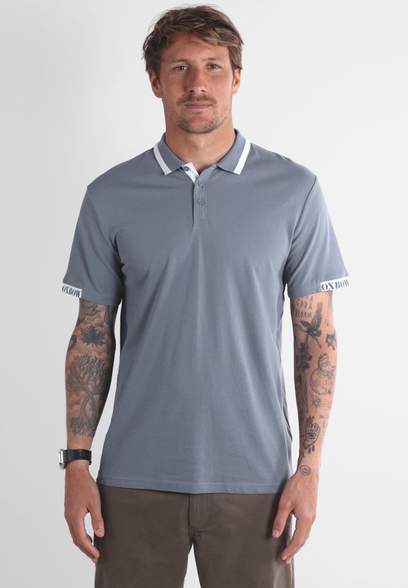 Un homme avec des tatouages porte une chemise polo grise avec un col blanc et un pantalon marron, se tenant devant un fond clair.