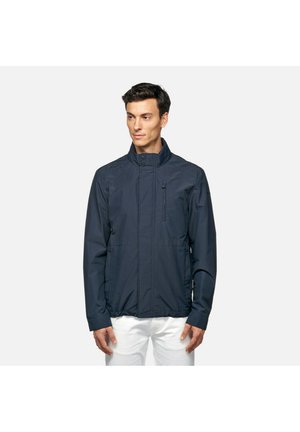 Giacca blu navy con colletto rialzato, chiusura frontale a zip, tasche laterali e tasca sul petto. Texture liscia, materiale leggero.