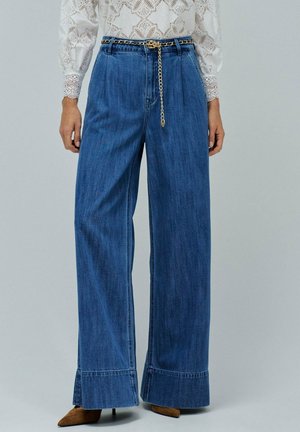 Femme portant un jean bleu taille haute et large avec une ceinture en chaîne dorée, un blouse en dentelle blanche et des escarpins bruns à bout pointu se tenant sur un fond uni.