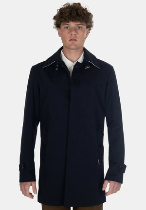 Giovane con capelli ricci che indossa un cappotto blu scuro sopra una camicia bianca con colletto, in piedi davanti a uno sfondo chiaro e semplice.