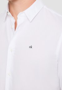 Camisa blanca de botones con cuello clásico, que presenta un pequeño logotipo "ck" bordado en el pecho. Textura de tela suave.