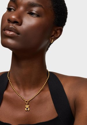 Goldkette mit einem Anhänger in Herzform und einem kleinen lila Edelstein. Das Model trägt große Ohrringe und ein schwarzes Oberteil.