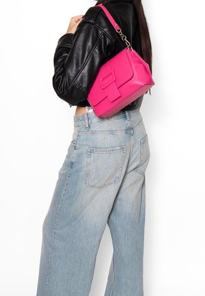 Vrouw in lichtblauwe jeans met wijde pijpen en een zwart leren jack, met een felroze schoudertas aan haar rechterkant.