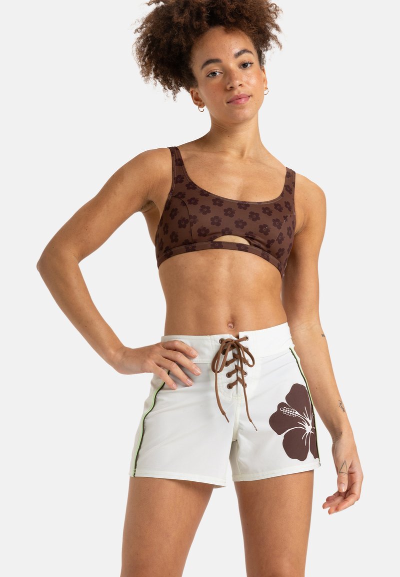 Hnědý květinový crop top s výřezem, spárovaný s bílými šortkami s hnědým potiskem ibišku a hnědým šněrováním vpředu.