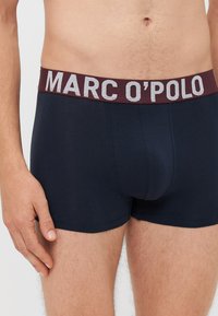 Tengerészkék boxer alsónadrág bordó elasztikus derékpánttal, amelyen ezüst betűkkel szerepel a "MARC O'POLO" felirat. Sima anyag, testhezálló fazon.
