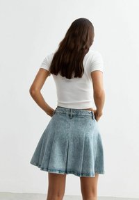 Hellblauer Jeansrock mit Falten, hoher Taille und ausgefranstem Saum. Kombiniert mit einem figurbetonten weißen kurzärmeligen Oberteil, von hinten betrachtet.