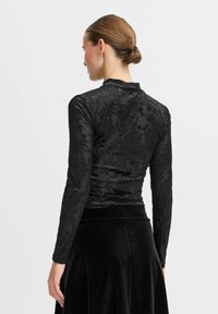 Top nero a maniche lunghe con texture floreale, colletto alto e silhouette aderente. Indossato con una gonna in velluto nero, che mette in mostra un tessuto liscio e morbido.