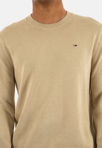 Maglione beige a maglia con scollatura a coste e maniche lunghe. Presenta un piccolo logo con accenti rossi, bianchi e blu sul petto.