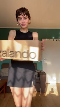 Caja de cartón marrón con "zalando" impreso en negro. Persona con una camiseta sin mangas negra y falda sostiene la caja, con un suelo de madera clara y una pared verde detrás.