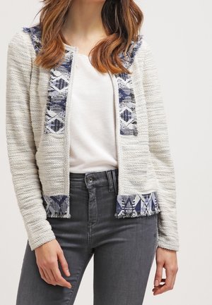 Witte textuur openvallende cardigan met blauwe geometrische patroonbies, gedragen over een wit shirt en gecombineerd met donkergrijze spijkerbroek.