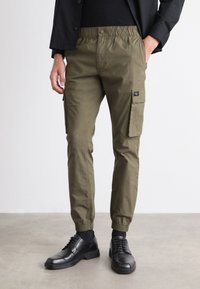 Pantalon cargo vert olive avec une taille élastique, une coupe effilée, des poches latérales et un petit patch de marque. Associé à des chaussures habillées noires.
