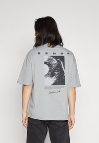 Una persona con cabello largo y oscuro lleva una camiseta gris claro con un gráfico abstracto en blanco y negro y el texto "SENSE" en la parte de atrás, combinada con pantalones negros.