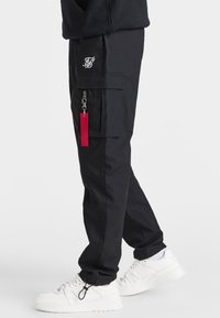 Svarta cargo-byxor med flera fickor, en röd tygskylt och en logopatch. Bärs med vita sneakers som visar en avslappnad passform.