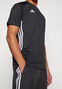 Svart träningsskjorta med korta ärmar, med vita Adidas-ränder på sidorna och meshdetaljer på övre framsidan.