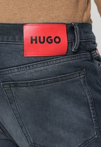 Jeans negri din denim cu o etichetă din piele roșie marcată „HUGO” la talie, având buzunare spate și detalii contrastante la cusături.