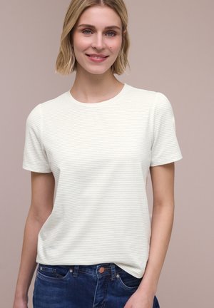 Kvinde med kort blondt hår iført en hvid kortærmet stribet t-shirt og blå jeans, smilende mod en neutral baggrund.