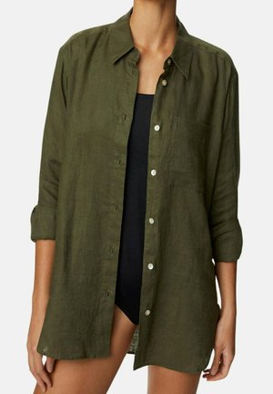 Personne portant une chemise longuement boutonnée couleur olive ouverte sur un haut noir sans manches ou un maillot de bain, avec les manches retroussées.