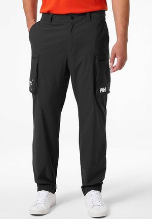 Man wearing black cargo pants with elastic cuffs, white sneakers, and an orange shirt, standing with hands at sides.