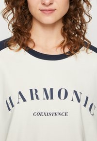 Cremfarbendes T-Shirt mit marineblauen Akzenten; mit dem Text "HARMONIC" und "COEXISTENCE" darunter, rundem Ausschnitt und kurzen Ärmeln.