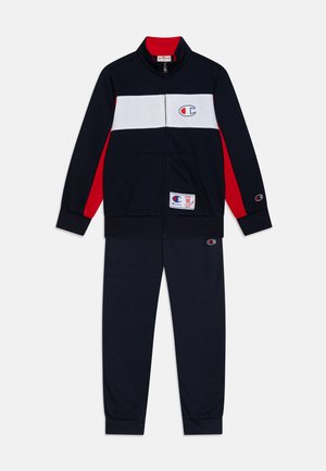 Chándal de color marino con chaqueta con cremallera que presenta franjas horizontales en blanco y rojo. Los pantalones son del mismo color marino. Incluye detalle de logo y bolsillos.