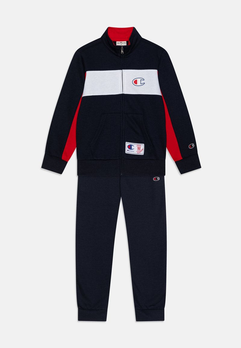 Marine blazer met ritssluiting, voorzien van witte en rode horizontale strepen. De broek is passend in marineblauw. Inclusief logo-detail en zakken.