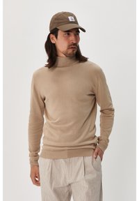 Pulover bej cu gât, realizat dintr-un material moale, cu mâneci lungi și un design ajustat, combinat cu pantaloni cu dungi deschise și o șapcă maro.