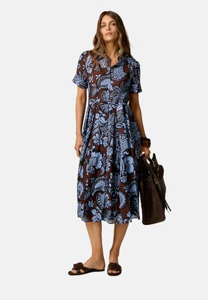 Vrouw die een bruine en blauwe bloemenjurk met korte mouwen draagt, een grote bruine gestructureerde handtas vasthoudt en bruine slides sandalen draagt.