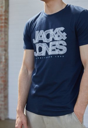 Homme portant un T-shirt bleu marine avec le texte "JACK & JONES Established 1990", debout, une main dans la poche d'un pantalon beige.