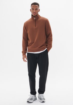 Sudadera marrón con cremallera en el cuello y mangas largas, combinada con pantalones negros y zapatillas grises, con un ajuste casual y un diseño minimalista.