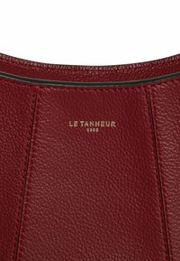 Borsa a mano in pelle rossa con texture a pelle di pesca, dotata di logo dorato in rilievo "LE TANNEUR 1898" e cuciture in contrasto lungo le cuciture.