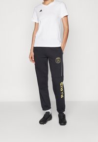 Biała koszulka z krótkim rękawem z czarnym logo, zestawiona z czarnymi spodniami dresowymi z żółtym napisem "PARIS" i okrągłym logo na lewej udzie.