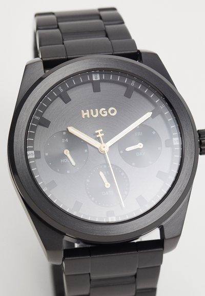 Montre noire en métal avec un boîtier rond, finition mate, et un cadran dégradé gris. Dotée d'accents dorés, d'un affichage de la date et de trois sous-cadrans.