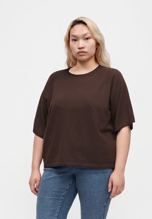 Ung kvinde med langt blondt hår iført en brun kortærmet top og blå jeans, stående foran en ensfarvet lys baggrund.