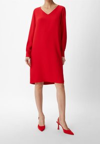 Vestido rojo de cuello en V con mangas largas y fruncidas y corte recto. Combinado con tacones rojos de puntera puntiaguda, sobre un fondo blanco.