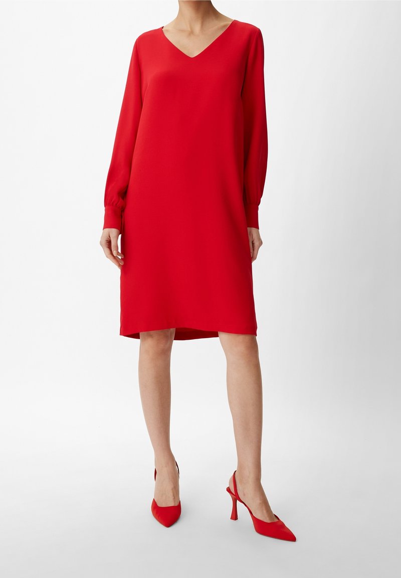 Vestido rojo de cuello en V con mangas largas y fruncidas y corte recto. Combinado con tacones rojos de puntera puntiaguda, sobre un fondo blanco.