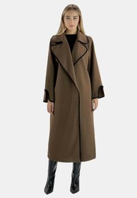 Impermeable largo marrón con ribete negro, solapas profundas y mangas amplias, hecho de tejido texturizado. Combinado con botas negras hasta la rodilla.