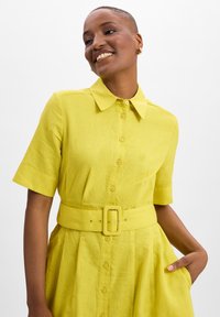 Yellow button-up dress with a collar, short sleeves, a belted waist, and pockets. Made from a textured fabric with a subtle sheen.