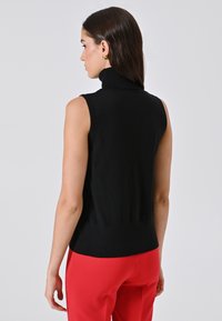Zwarte mouwloze turtleneck top met een fitted taille en geribbelde textuur, gedragen met rode broek. Uitzicht van achteren, dat de nekdetail laat zien.