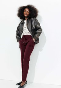 Veste bomber en cuir noir, blouse blanche à motifs, pantalon bordeaux à cordon de serrage et ballerines noires à pois blancs. Textures lisses.