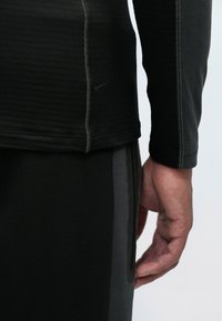 Čierny atletický top s dlhým rukávom z textúrovanej tkaniny, s logom Nike. Spárovaný s čiernymi nohavicami, ktoré majú jemné šedé bočné akcenty.