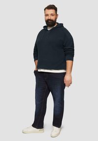 Marineblaues Ripp-Hoodie mit Kordelzug, über einem hellen Hemd getragen, kombiniert mit dunklen Jeans und weißen Sneakers. Vor einem grauen Hintergrund präsentiert.