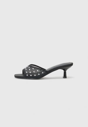 Sandal de abertura frontal preta com um salto baixo, apresentando um tecido transparente adornado com cravos claros decorativos e uma sola lisa.