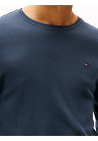 Granatowy sweter bawełniany z okrągłym dekoltem o gładkiej fakturze, z małą naszywką z logo w kolorach czerwonym, białym i niebieskim na lewej piersi.