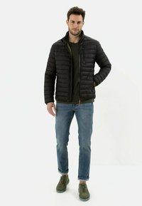 Sort pufferjakke med lynlåsåbning, høj krave og quiltet mønster, parret med en mørkegrøn skjorte, blå jeans og grønne sneakers.