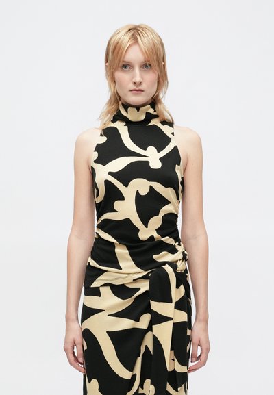 Donna con capelli biondi che indossa un abito nero senza maniche a collo alto con grandi motivi floreali astratti beige, in piedi su uno sfondo semplice.