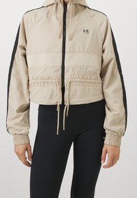 Beskarvet beige jakke med sorte sidelapper, snøre i livet og hætte. Har elastiske manchetter og et logo på venstre bryst.